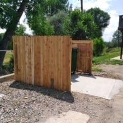 Wooden fence enclosure for a trash bin in a park setting with green trees.
