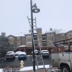 Snowy scene of a ladder next to a light pole at an apartment complex parking lot.