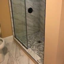Modern shower with glass enclosure, marble walls, and pebble stone floor in a bathroom setting.