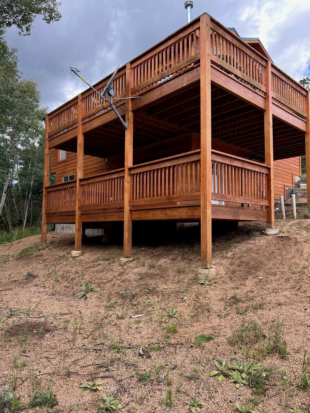 Two-story wooden cabin with balconies, surrounded by sandy landscape and greenery.
