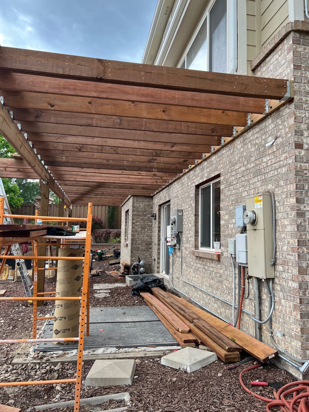 Wooden patio cover under construction next to a brick home with electrical utilities visible.