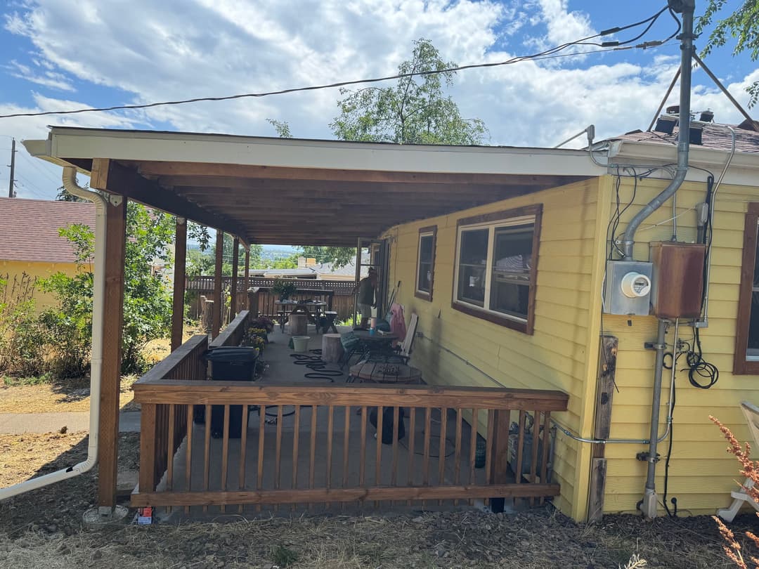 Yellow house with porch and wooden railing, surrounded by green plants and blue sky.